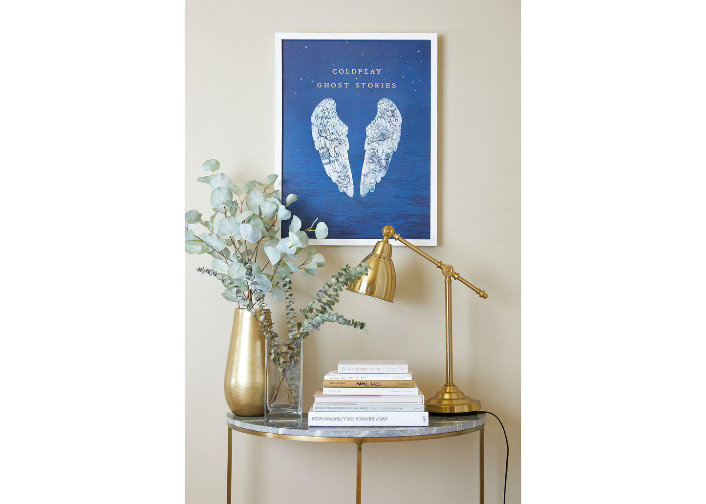 Ghost Stories lithograph on a white frame, hung on a beige wall, above a small table shaped like a crescent moon, with a marble top and golden legs. The table has golden and clear vases with eucalyptus, books and a golden desk lamp. The white angel wings add a touch of calm and serenity, complementing the room’s elegant decor
