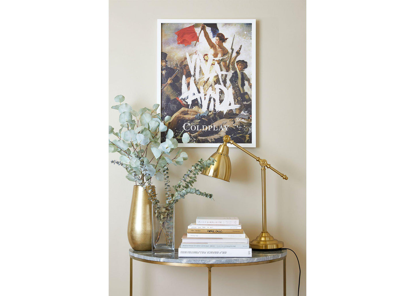 Viva La Vida Lithograph on a white frame, hung on a beige wall, above a small table shaped like a crescent moon, with a marble top and golden legs. The table has golden and clear vases with eucalyptus, books and a golden desk lamp. The painting’s references to art and music give the room a dynamic, cultured feel