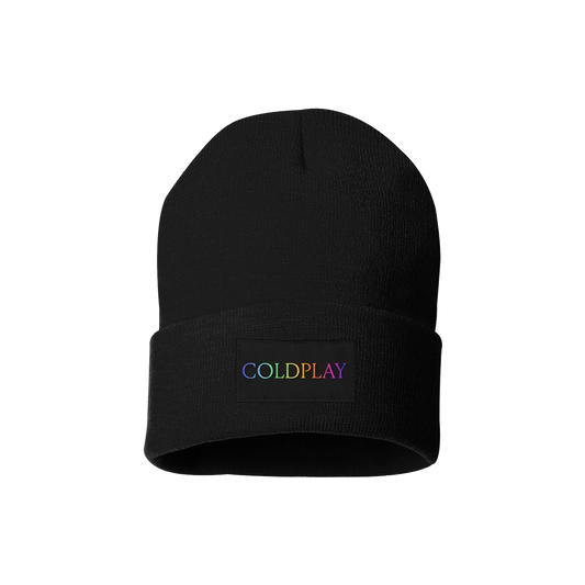 Black knitted winter beanie, with the word COLDPLAY written on the front, in rainbow gradient letters. C is lilac, O is light blue, L is green, D green to yellow, P is orange, L dark orange, A is pink fuchsia, and Y is lilac. The colors blend smoothly from one letter to the next. This logo is on a small rectangular patch, sown onto the fabric. The beanie looks very cozy&has a folded brim. The design is minimalistic and modern