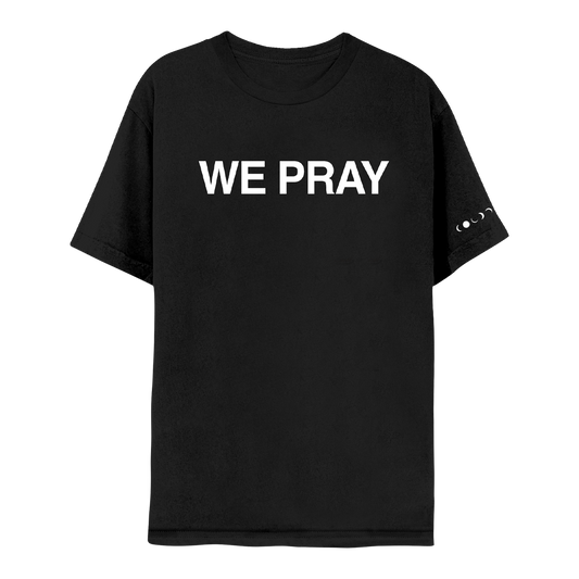 Front: WE PRAY is printed in large, white, bold letters across the chest of a black short sleeve t-shirt. Coldplay is spelled out in white phases of the moon at the hem of the left sleeve.